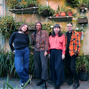 4 people looking at camera, wearing bright lovely clothes, before a plethora of potted plants